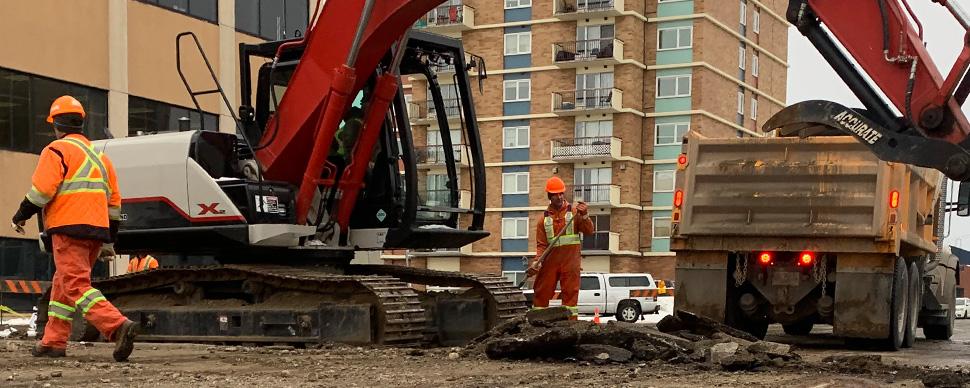 Excavator and dump truck in construction site, with two workers wearing hardhats and orange high visibility clothing.